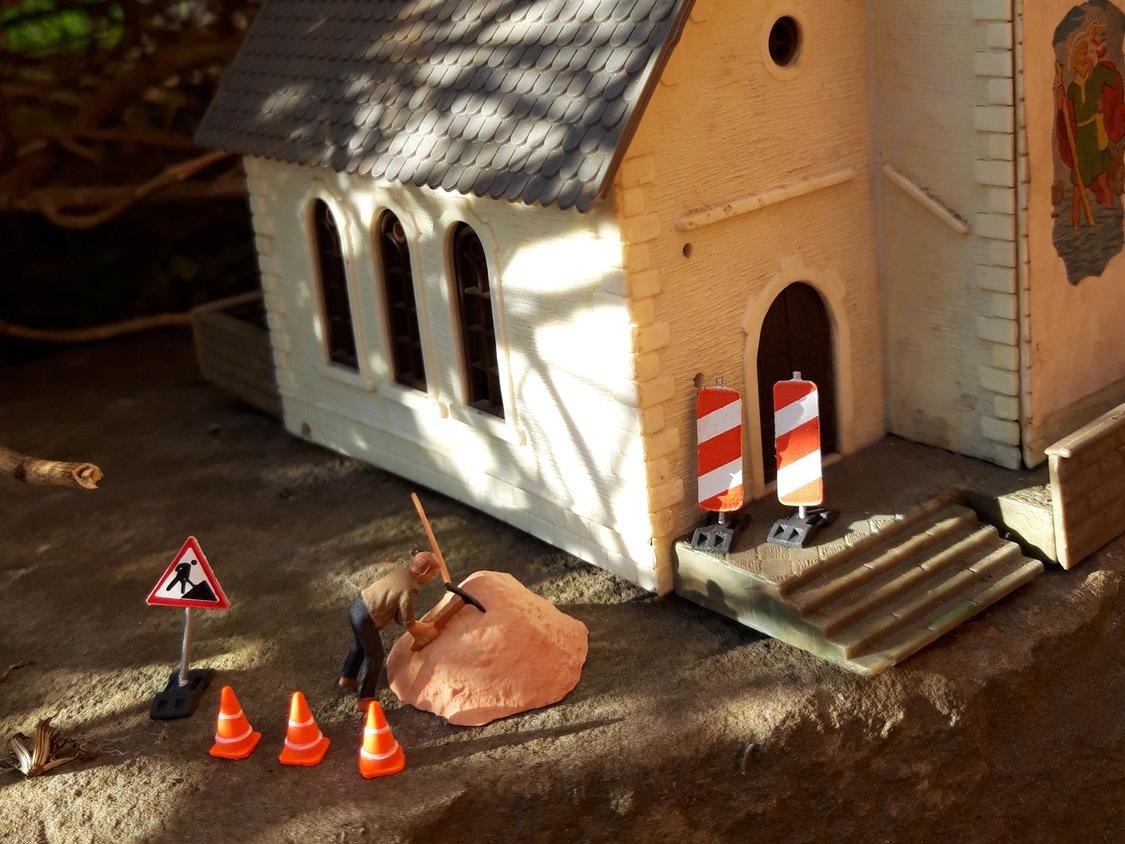 Vor einem Kirchengebäude im leichten Sonnenlicht ist eine Baustelle eingerichtet, ein Bauarbeiter arbeitet an einem Hügel mit Sand.