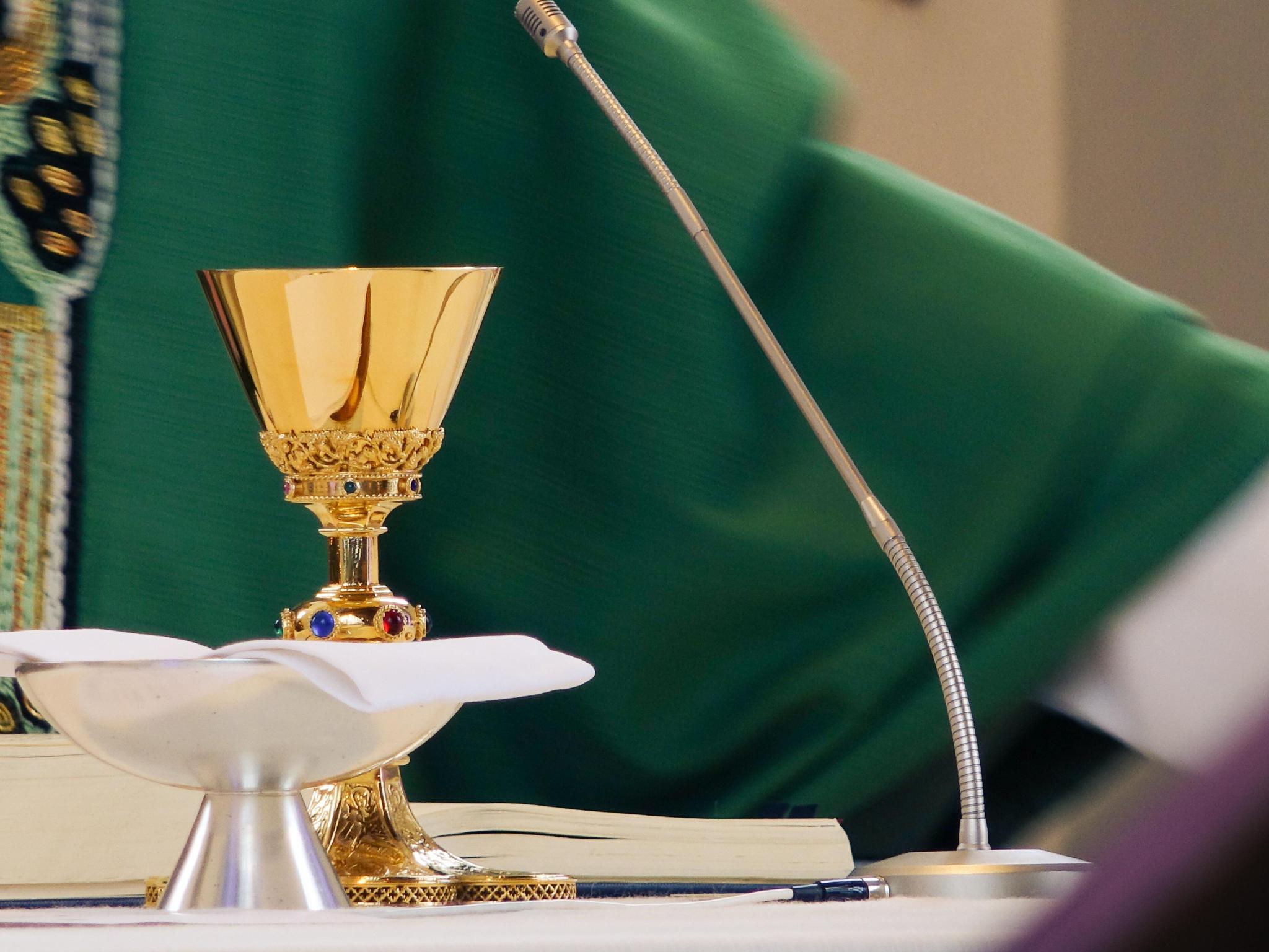 Auf dem Altar stehen Kelch und Patene, dahinter sieht man einen Teil eines Priesters im grünen Messgewand.