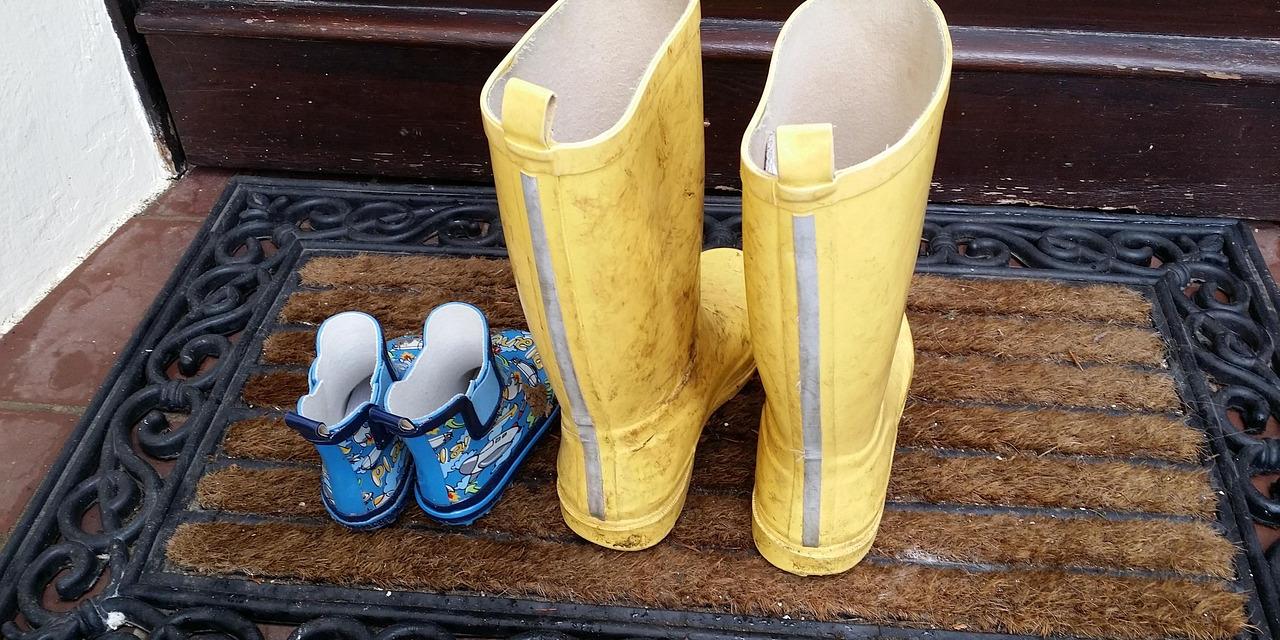 Nebeneinander auf einer Fußmatte steht ein Paar Gummistiefel eines Kindes (blau) neben einem Paar für Erwachsene (gelb).